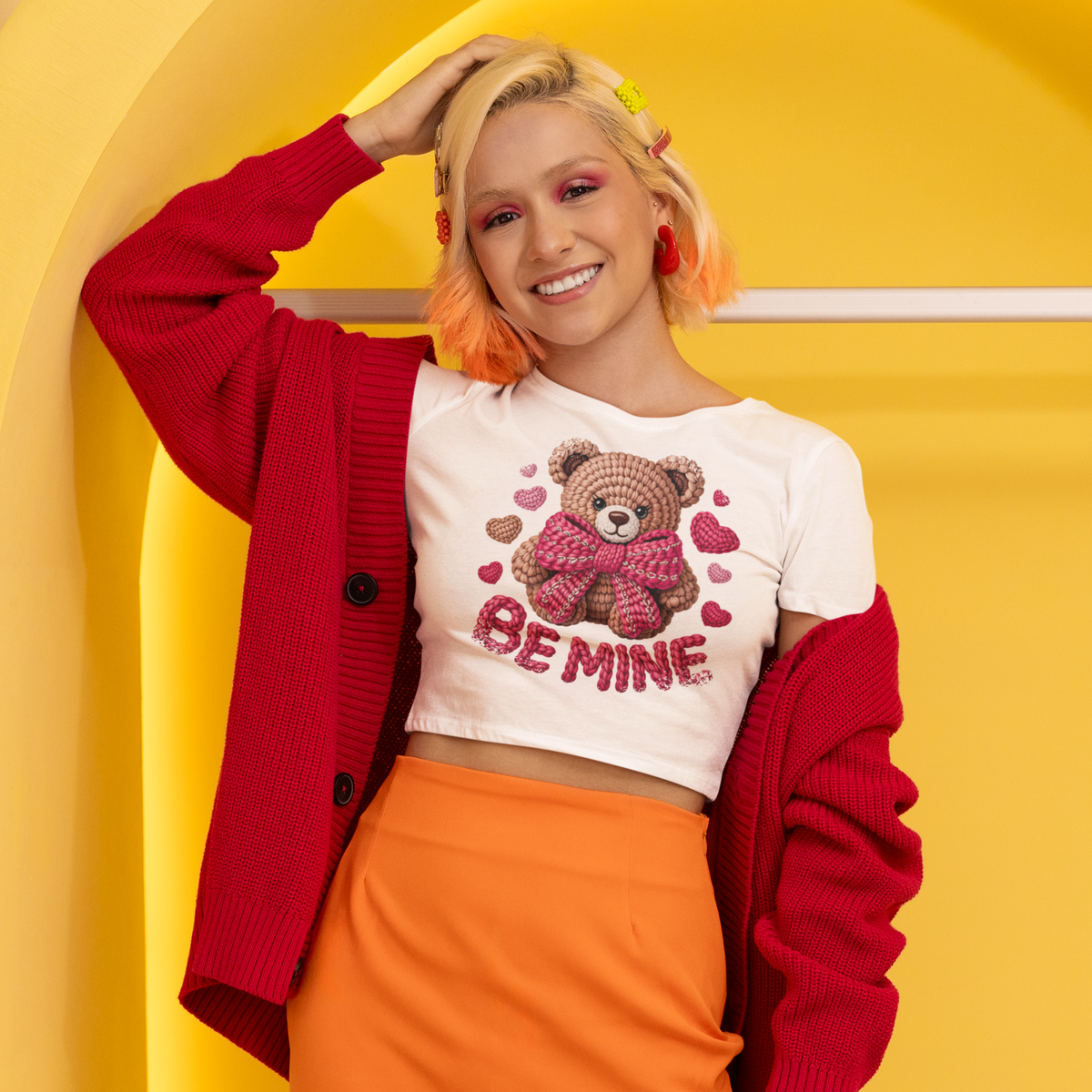 Person wearing a white shirt with a teddy bear graphic and 'Be Mine' text, red cardigan, and orange pants against a yellow background.