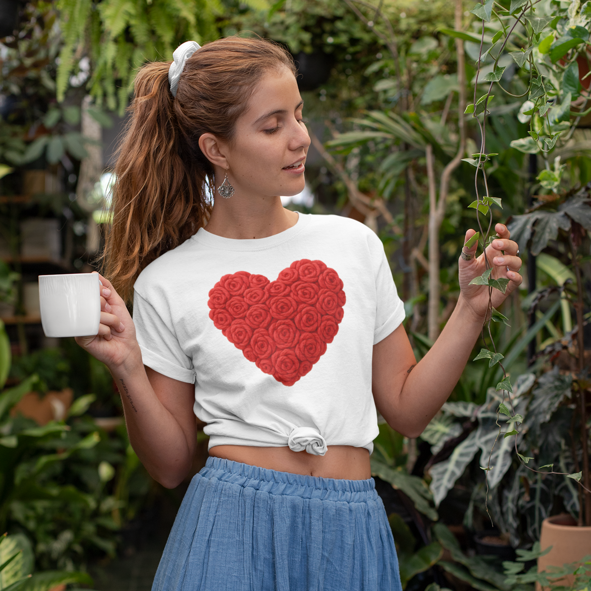 Woman wearing a white t-shirt with a red heart design, holding a cup in a garden setting.